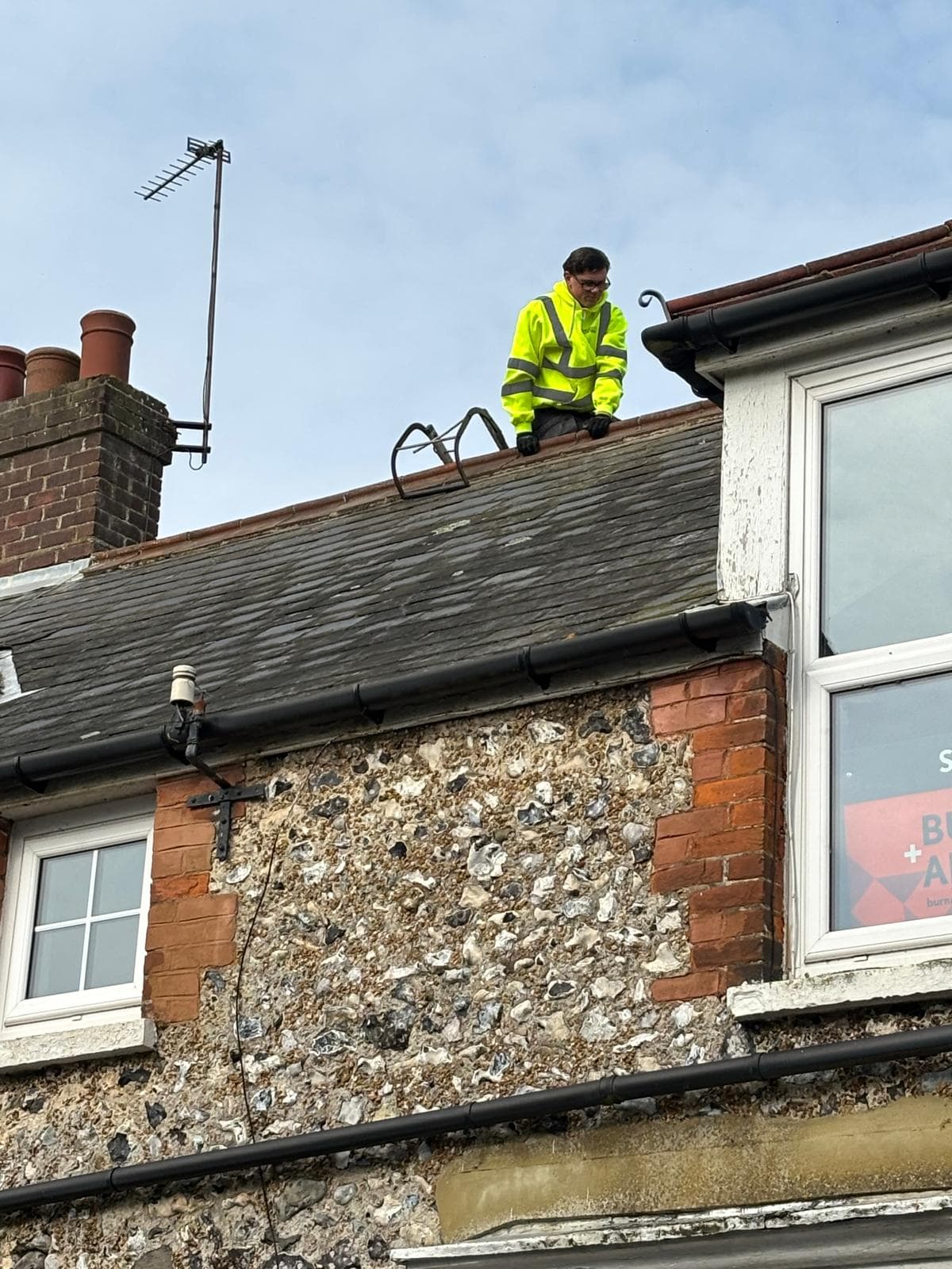 A roofer on the roof of a flint and brick house, above its gutter and fascia