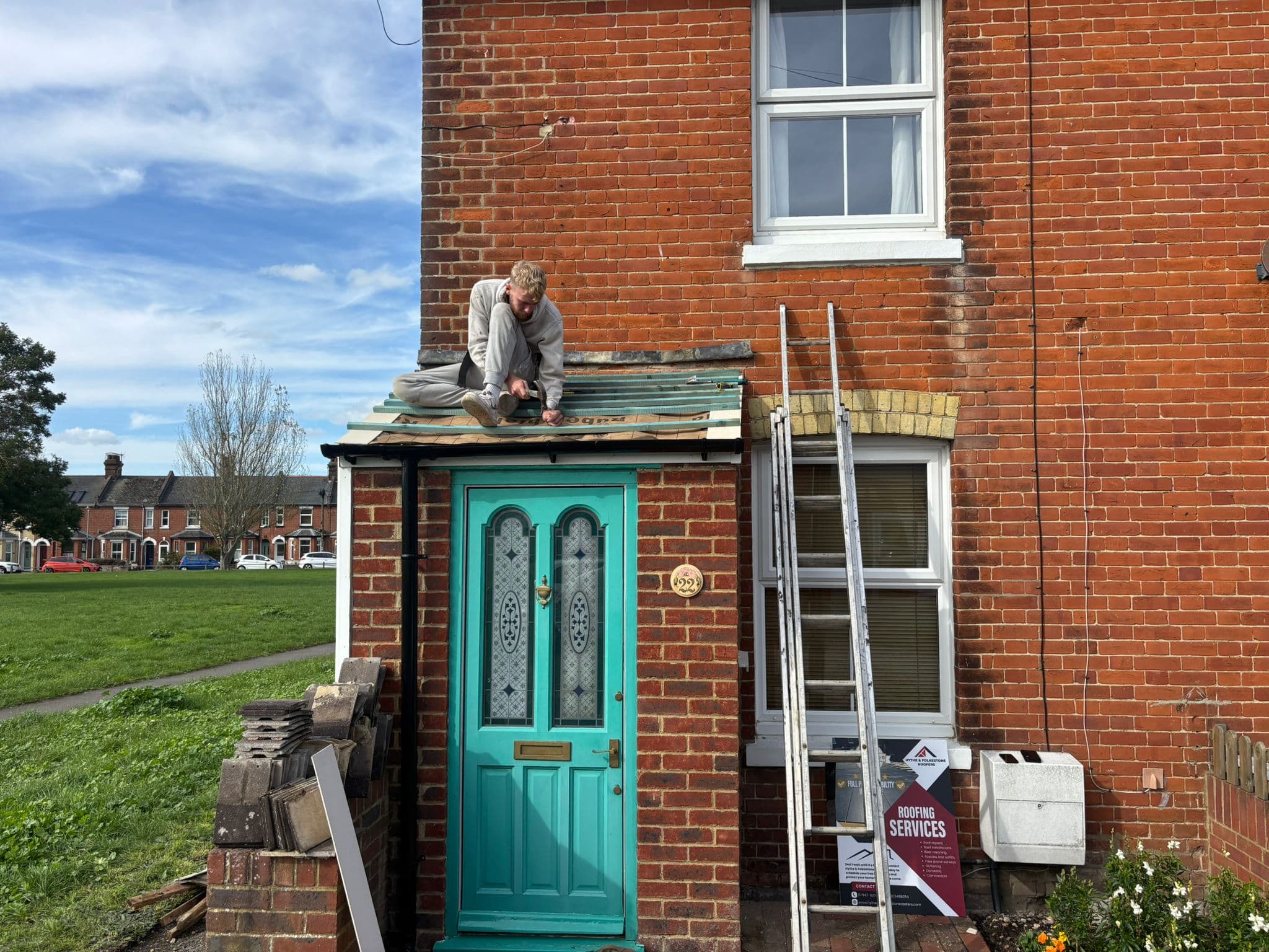 A roofer at work on a small porch roof above a teal front door