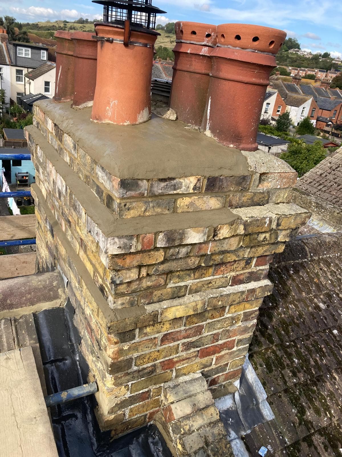 A brick chimney stack with fresh mortar flaunching around its clay pots and new flashing at its base, repaired from a scaffold