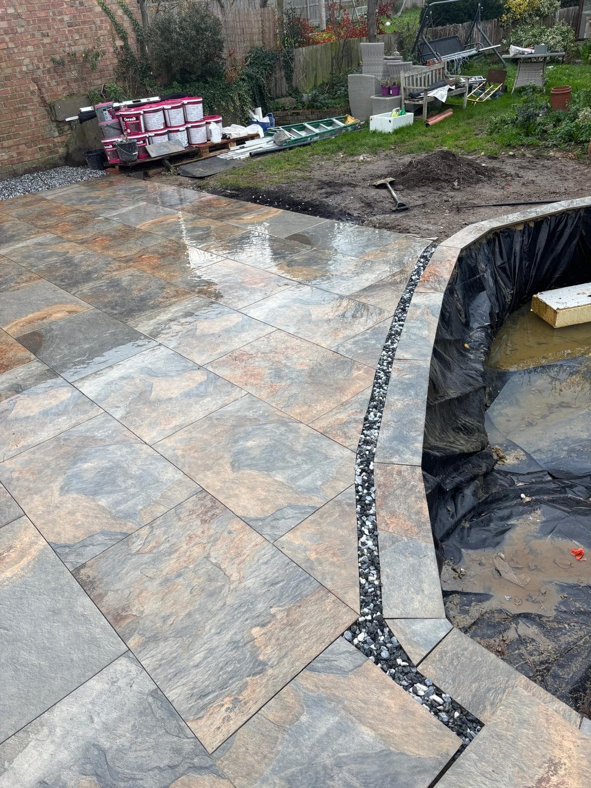 A newly laid stone effect patio with a curved edge beside a garden pond under construction